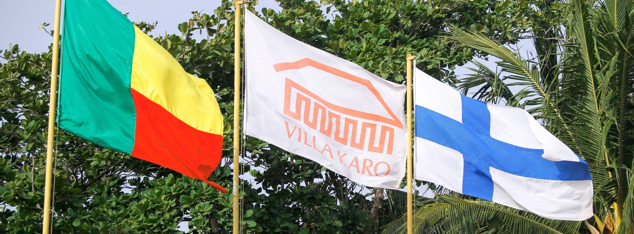 Kolme lipputankoa, joissa on Beninin, Villa Karon ja Suomen lippu. Vasemmalla: vihreää, punaista ja keltaista. Keskellä: valkoista ja oranssia. Oikealla: Sininen risti valkoisella taustalla.